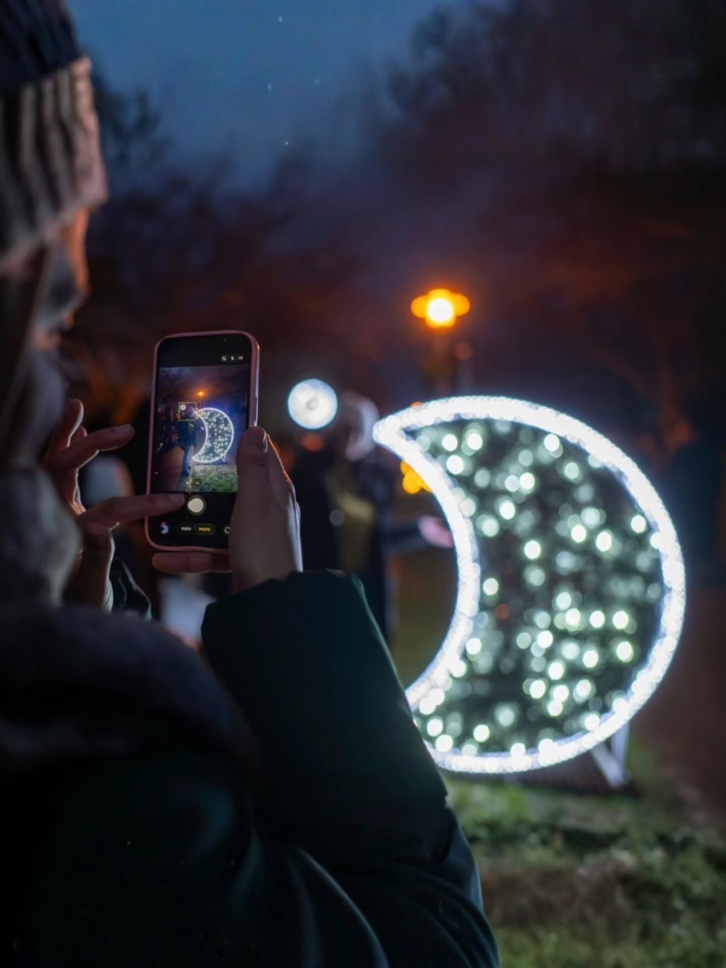 Dieser Alternativtext wurde KI-generiert: Eine Person hält ein Smartphone vor sich und fotografiert eine beleuchtete Mond-Installation. Im Hintergrund sind weitere Lichter und verschwommene Gestalten zu sehen.