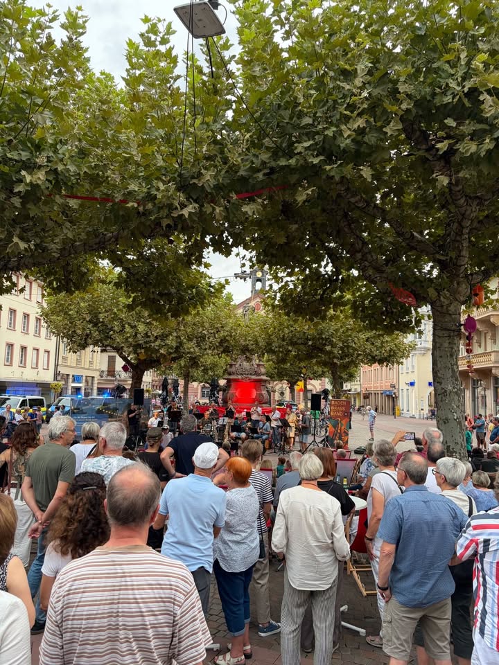 Eine Menschenmenge steht auf einem Platz und schaut auf eine Bühne, auf der die "Baden Blues Band" auftritt. Große Bäume hängen über dem Platz, im Hintergrund sind Gebäude und ein Brunnen zu sehen. (KI-Alternativtext)