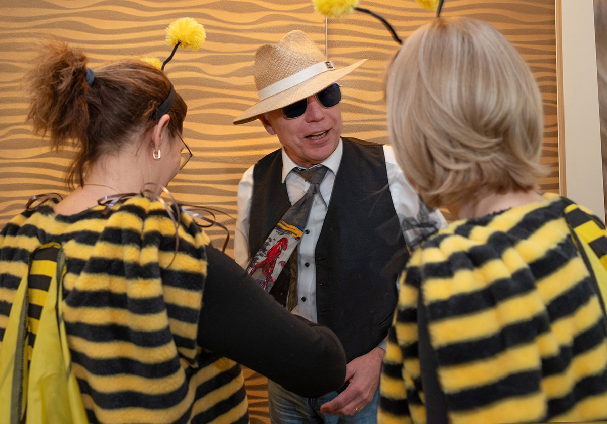 Dieser Alternativtext wurde KI-generiert: Mehrere Personen stehen zusammen und unterhalten sich. Einige tragen Hüte und Kleidung im Bienen-Design.