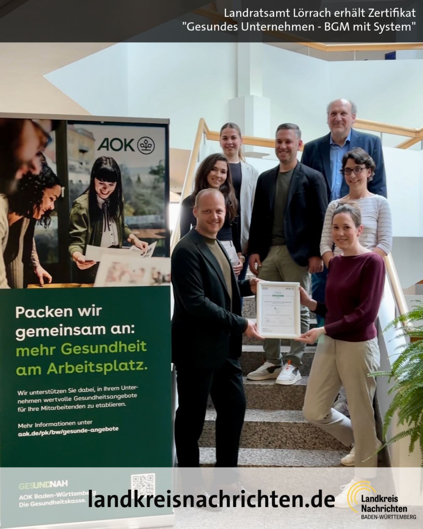 Dieser Alternativtext wurde KI-generiert: Farbfoto zeigt eine Gruppe von Menschen, die vor einem Plakat und einer Glasfassade stehen. Ein Zertifikat wird überreicht, während im Hintergrund Informationen zu Gesundheit am Arbeitsplatz gezeigt werden.