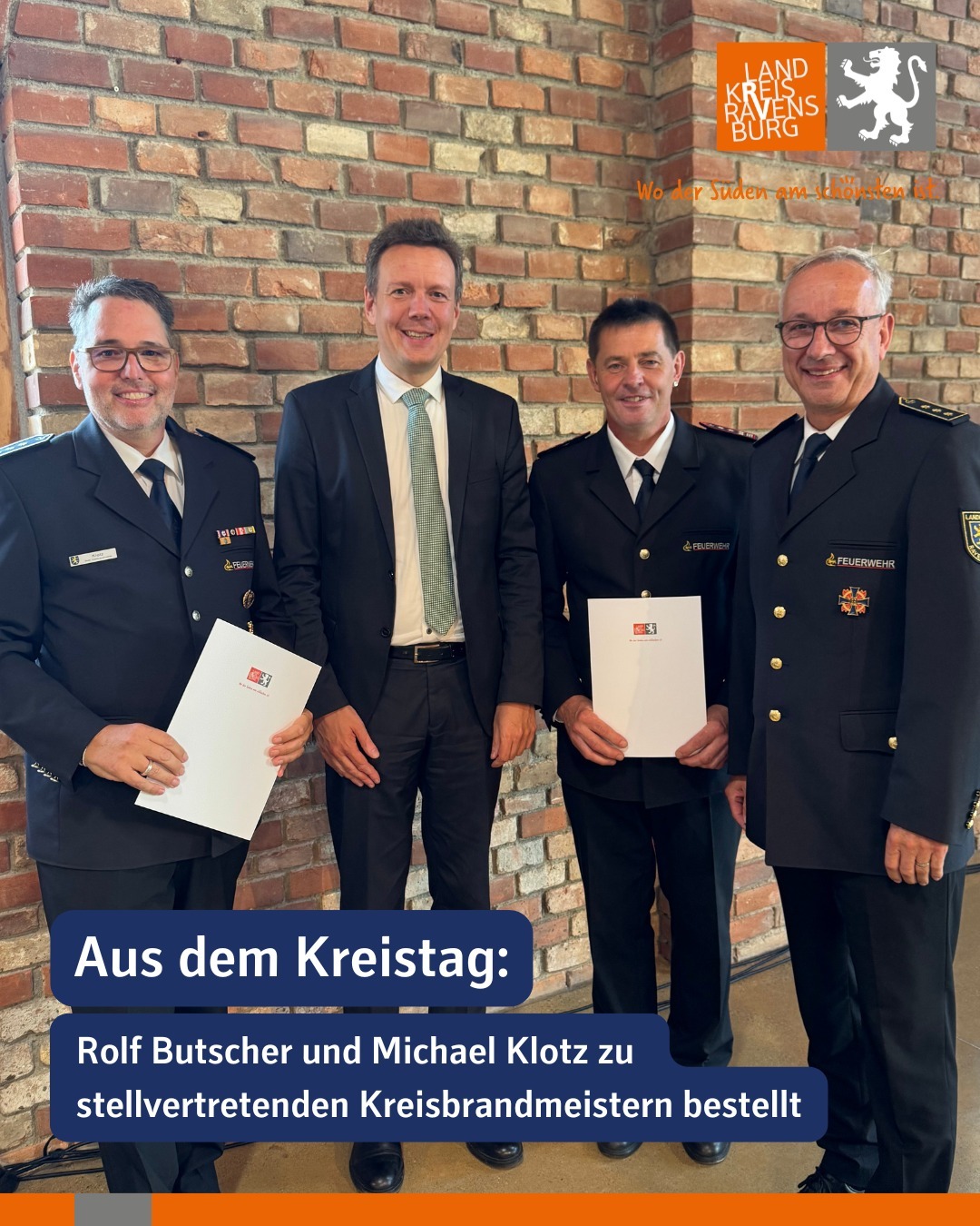 Vier Personen stehen vor einer Ziegelwand. Einige der Personen tragen Uniformen und halten Dokumente.