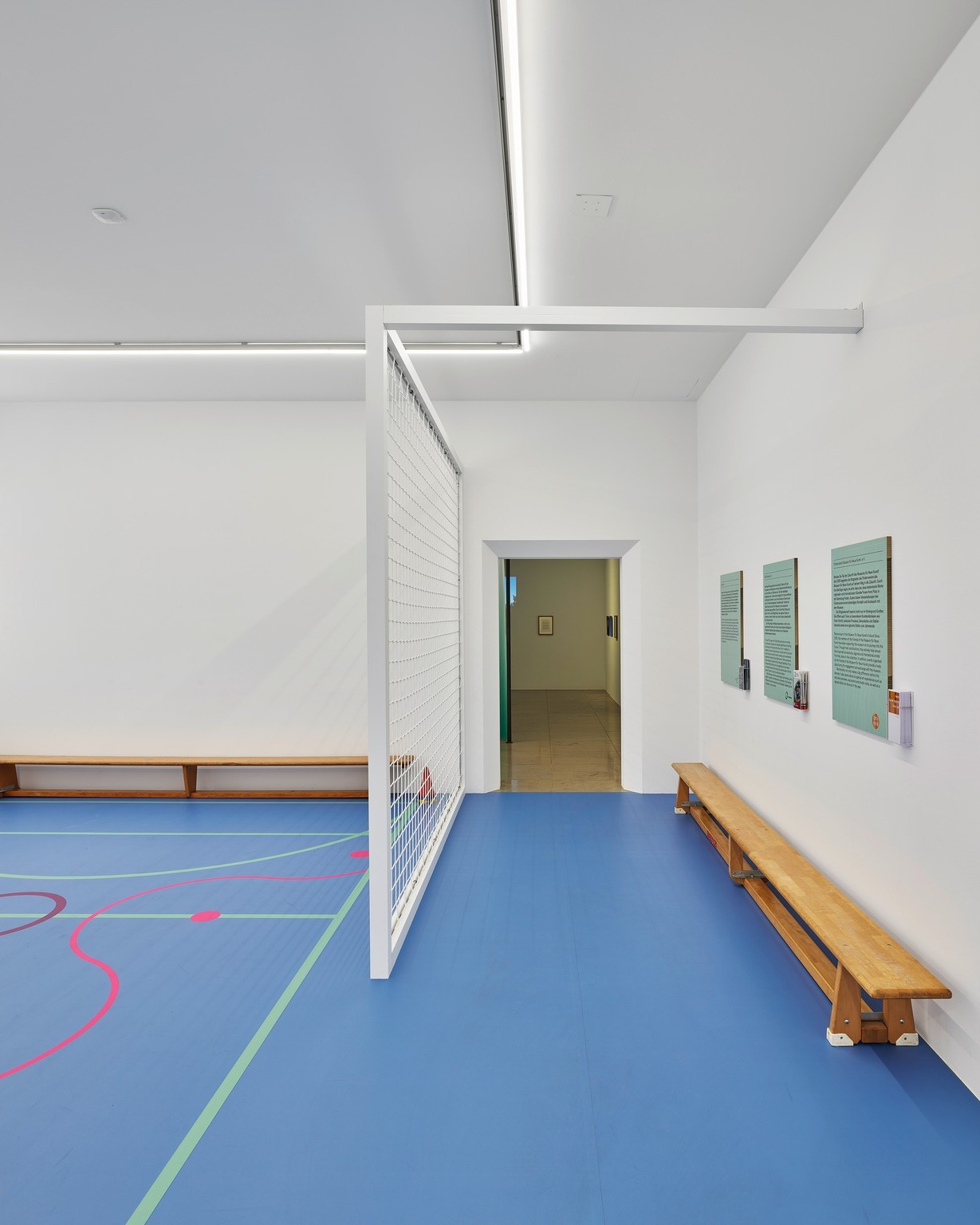 Heller Raum mit blauem Boden und farbigen Linien, geteilt durch eine weiße Trennwand mit Gitterstruktur. An der rechten Wand stehen eine lange Holzbank und drei grüne Informationsplakate. Im Hintergrund führt eine Tür in einen weiteren Raum. (KI-Alternativtext)