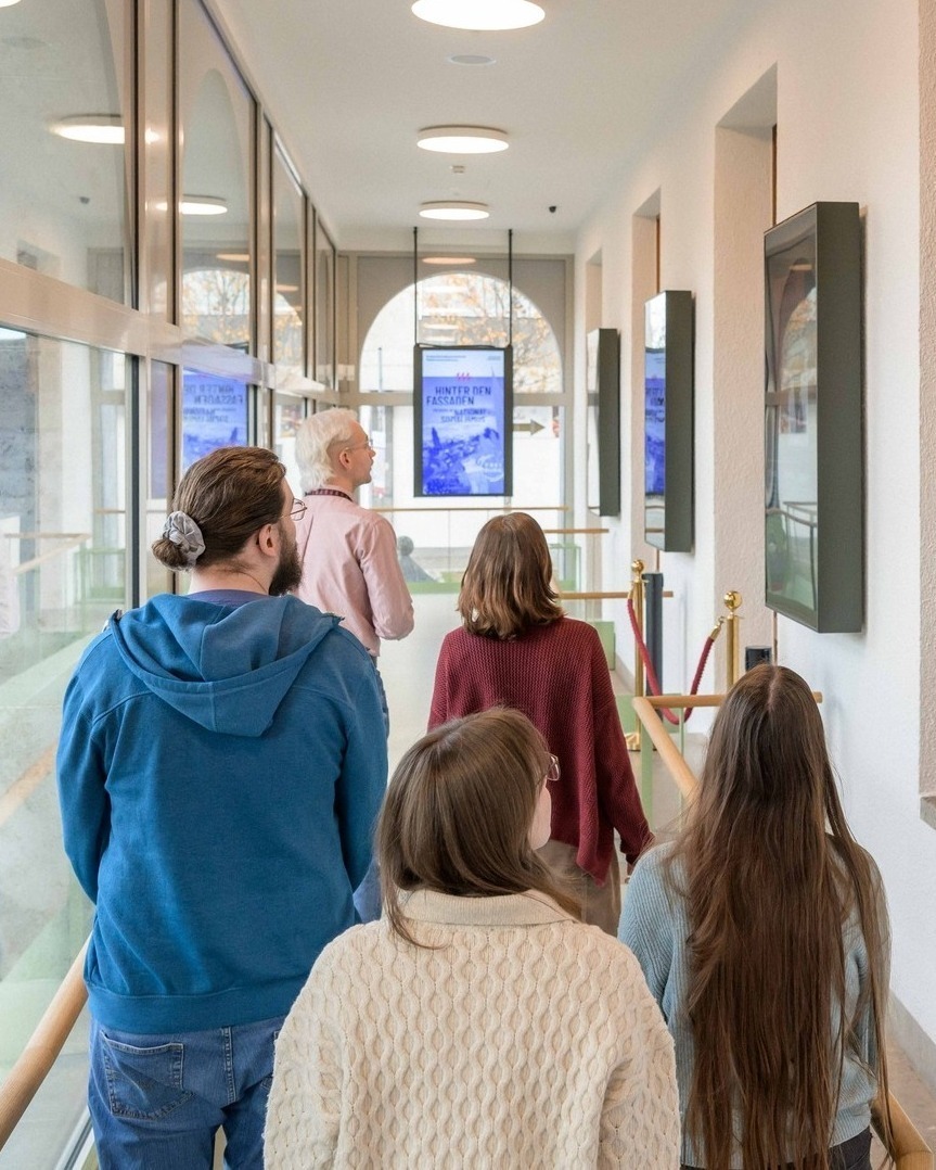 Von hinten gesehen geht eine Gruppe von fünf Personen einen hellen Flur entlang, der durch rote Seile abgegrenzt ist. Sie blicken auf digitale Bildschirme, die an der Decke hängen und an der Wand montiert sind. Auf einem der Bildschirme steht der Text „HINTER DEN FASSADEN“. (KI-Alternativtext)
