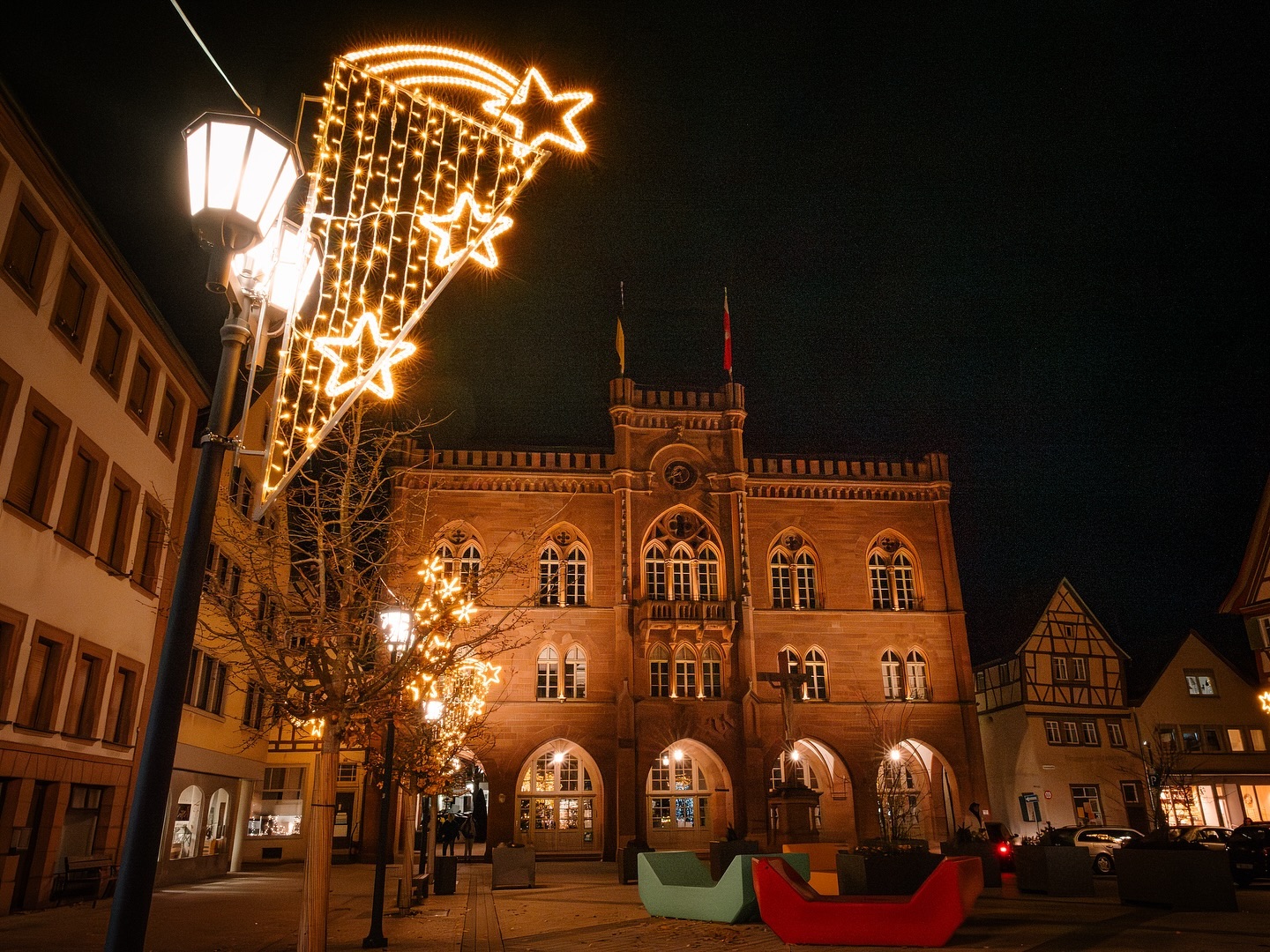 Festlich beleuchtetes Rathaus mit Sternen-Lichterketten an einem Abend. Im Vordergrund sind moderne Sitzgelegenheiten zu sehen.