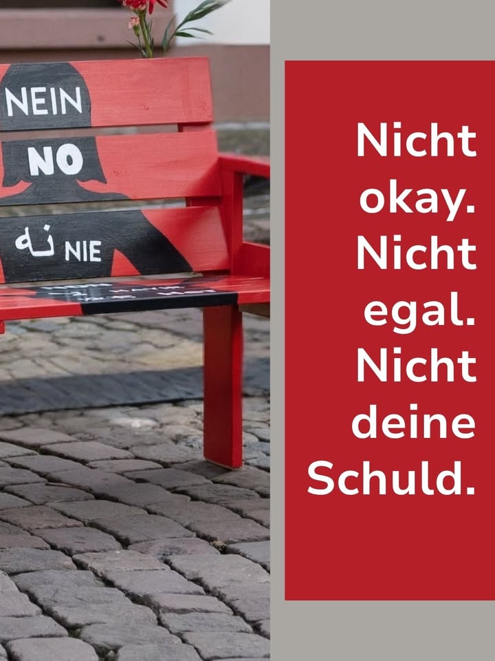 Eine rote Bank ist mit den Worten „NEIN“, „NO“ und „NIE“ bemalt. Rechts daneben steht auf rotem Grund in weißer Schrift: „Nicht okay. Nicht egal. Nicht deine Schuld.“ (KI-Alternativtext)