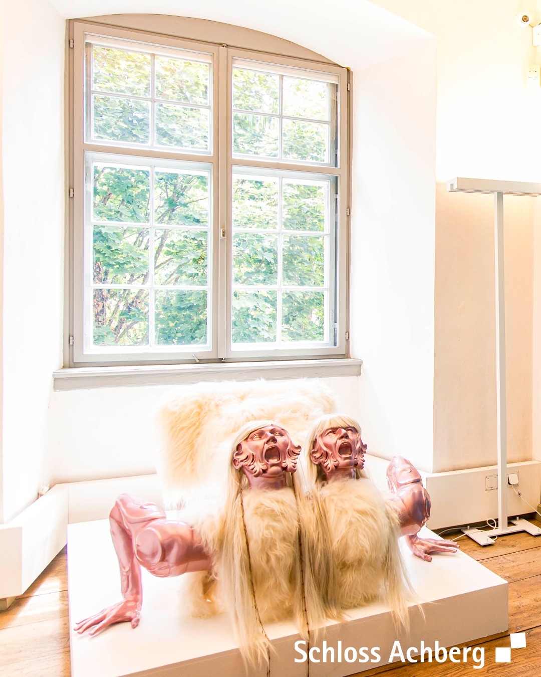 Dieser Alternativtext wurde KI-generiert: Ein Innenraum mit einem Fenster, durch das Bäume sichtbar sind. Im Vordergrund steht eine skulpturale Installation aus Fell und möglicherweise menschlichen Attributen auf einer weißen Plattform.