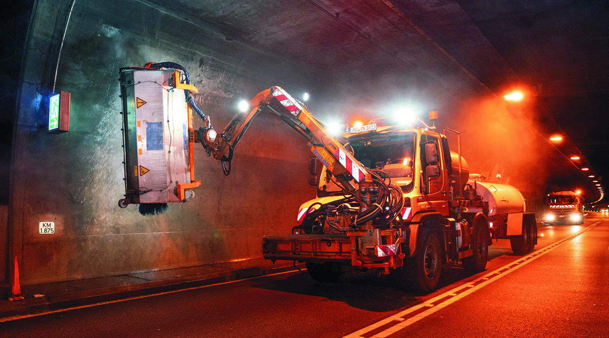 Dieser Alternativtext wurde KI-generiert: Ein Fahrzeug mit einem Kranarm steht in einem Tunnel. Im Hintergrund sind weitere Fahrzeuge und rote Warnlichter zu sehen.