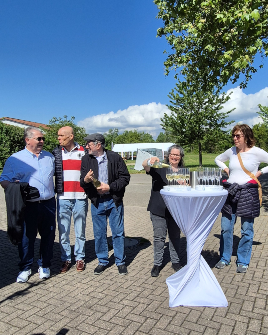Eine Person gießt draußen an einem weißen Stehtisch Sekt in Gläser, während vier weitere Personen daneben stehen und zuschauen. Im Hintergrund sind Bäume, ein Zelt und ein blauer Himmel mit Wolken zu sehen. (KI-Alternativtext)