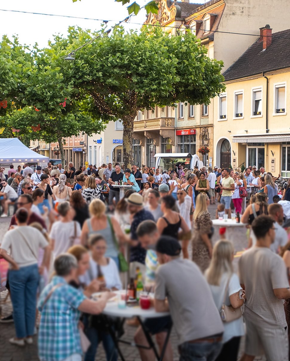 Auf einer belebten Straße findet ein Straßenfest statt, bei dem viele Personen stehen oder an weißen Tischen sitzen und trinken. Im Hintergrund stehen Gebäude mit Ladenschildern wie „KRU“ und „BOUTIQ“ sowie ein großer Baum mit Lichterketten. Links im Bild ist ein weißes Zelt zu sehen. (KI-Alternativtext)