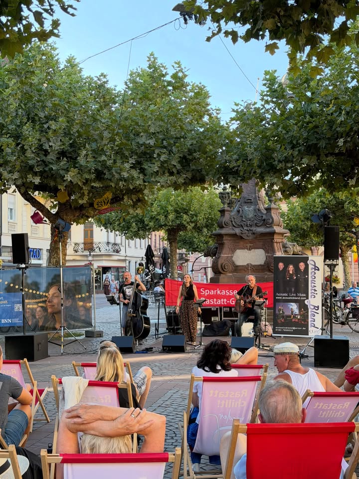 Drei Musiker spielen auf einer kleinen Bühne im Freien, während ein Publikum in Liegestühlen mit der Aufschrift "Rastatt Chillt!" zuschaut. Hinter der Band stehen ein rotes Banner mit der Aufschrift "Sparkasse Rastatt-G" und ein vertikales Banner für "Acoustic Blend". Im Hintergrund befindet sich ein großer Brunnen auf einem Platz mit Bäumen. (KI-Alternativtext)