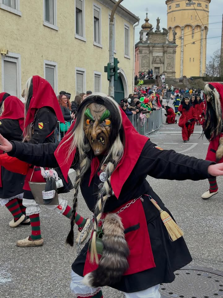 Dieser Alternativtext wurde KI-generiert: Eine farbenfrohe Aufnahme zeigt Figuren mit auffälligen Masken und Kostümen auf einer Straße. Im Hintergrund sind Menschen und Gebäude zu sehen, was auf eine Veranstaltung oder einen Umzug hindeutet.
