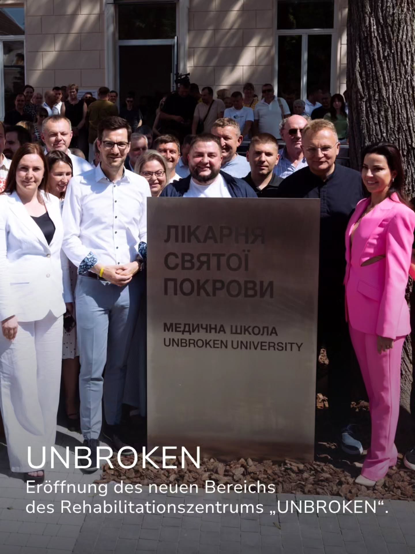 Eine Gruppe von Personen steht vor einem großen Schild mit der Aufschrift "ЛІКАРНЯ СВЯТОЇ ПОКРОВИ" und "МЕДИЧНА ШКОЛА UNBROKEN UNIVERSITY". Im Hintergrund ist eine weitere Menschenmenge vor einem Gebäude zu sehen. Ein Text-Overlay unten links lautet "UNBROKEN Eröffnung des neuen Bereichs des Rehabilitationszentrums „UNBROKEN“". (KI-Alternativtext)