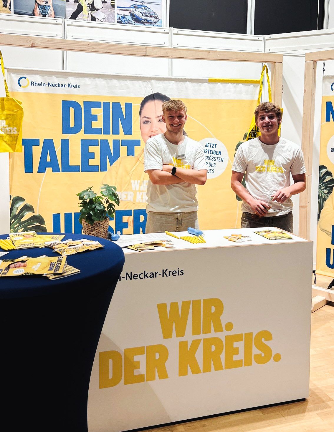 Zwei Personen stehen lächelnd hinter einem weißen Stand mit der Aufschrift „WIR. DER KREIS.“ und „Rhein-Neckar-Kreis“. Im Hintergrund hängt ein gelbes Banner mit dem Text „DEIN TALENT“. Auf der Theke und einem blauen Nebentisch liegen Flyer, gelbe Stifte und eine Pflanze. (KI-Alternativtext)