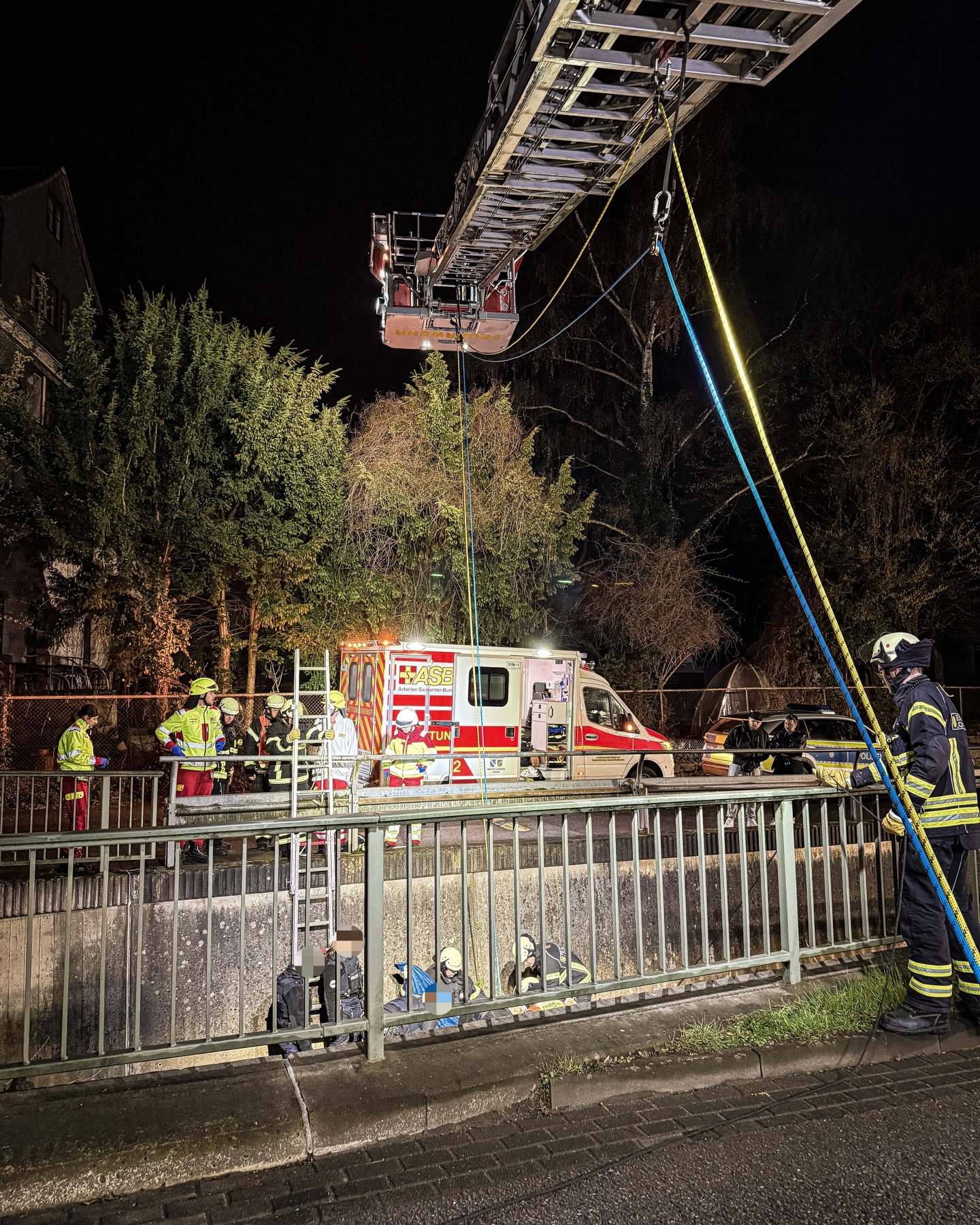 Bei Nacht findet ein Rettungseinsatz statt, bei dem eine Feuerwehrauslegerplattform hochgezogen ist und Seile herabhängen. Auf einer Brücke stehen Feuerwehrleute und ein Rettungswagen, während unten hinter einem Geländer Personen am Boden sitzen und von Helfern betreut werden. Im Hintergrund ist ein Polizeifahrzeug zu sehen. (KI-Alternativtext)
