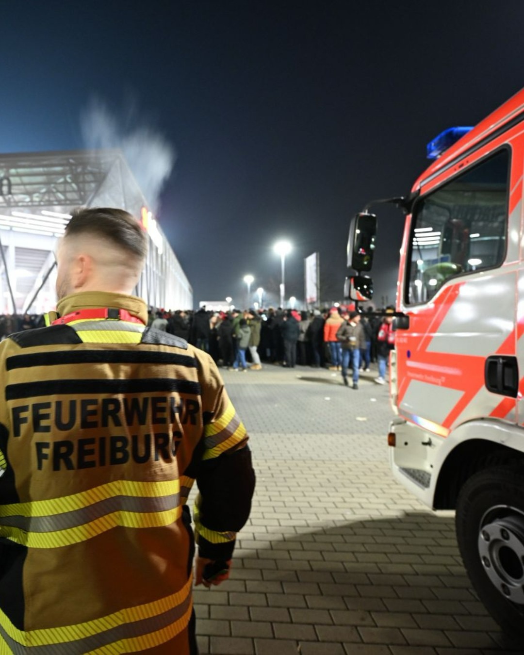 Dieser Alternativtext wurde KI-generiert: Ein Rettungsfahrzeug steht in der Nähe einer Menschenmenge. Im Vordergrund ist die Rückseite einer Person mit einer reflektierenden Jacke zu sehen.