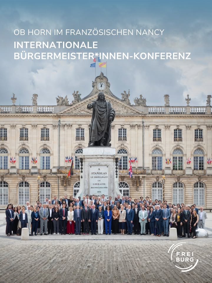 Gruppenfoto von Teilnehmenden der Internationalen Bürgermeister*innen-Konferenz vor einem historischen Gebäude in Nancy, Frankreich, mit einer Statue im Hintergrund und dem Logo von Freiburg unten rechts. (KI-Alternativtext)
