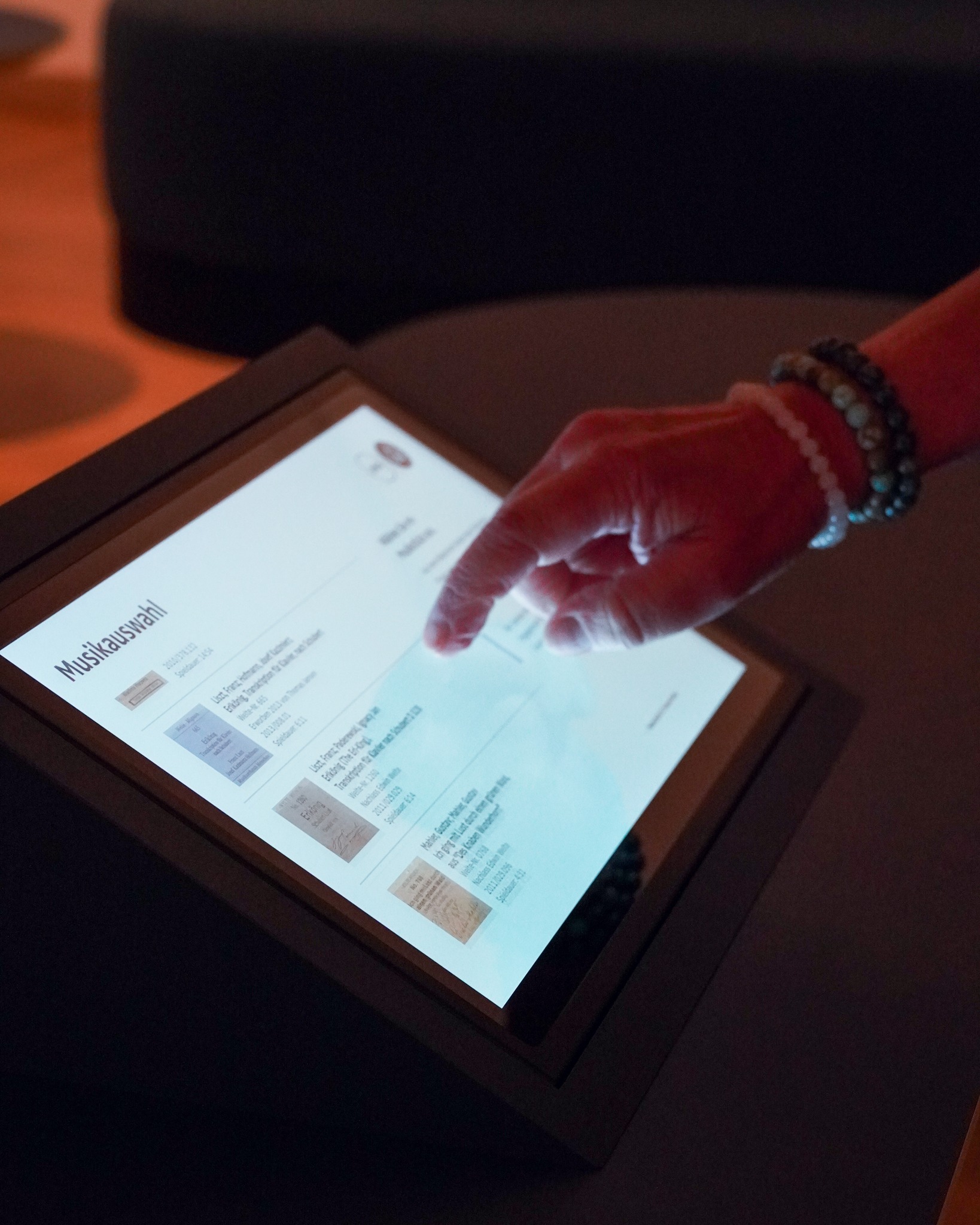 Eine Hand berührt einen Touchscreen mit der Überschrift "Musikauswahl". Auf dem Display ist eine Liste von Musiktiteln mit kleinen Vorschaubildern zu sehen. Der Zeigefinger tippt auf einen der Einträge. (KI-Alternativtext)