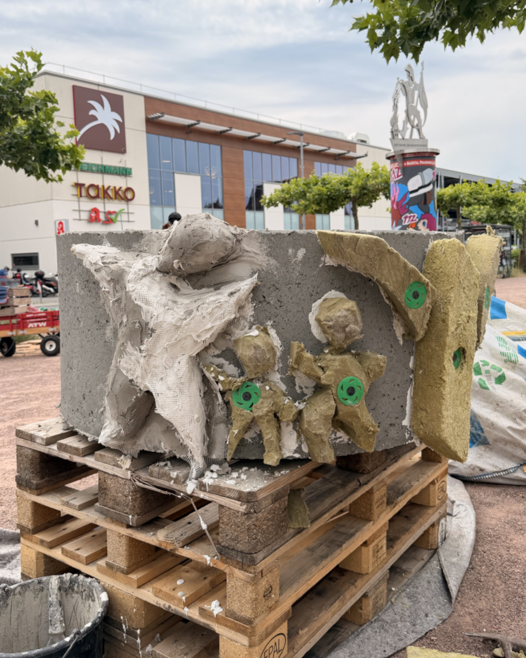 Skulptur aus Beton und Papiermasse mit zwei kindähnlichen Figuren und grünen Kreisen, auf Holzpaletten vor einem Einkaufszentrum mit Schildern wie „TOKKO“ und einer Statue im Hintergrund. (KI-Alternativtext)