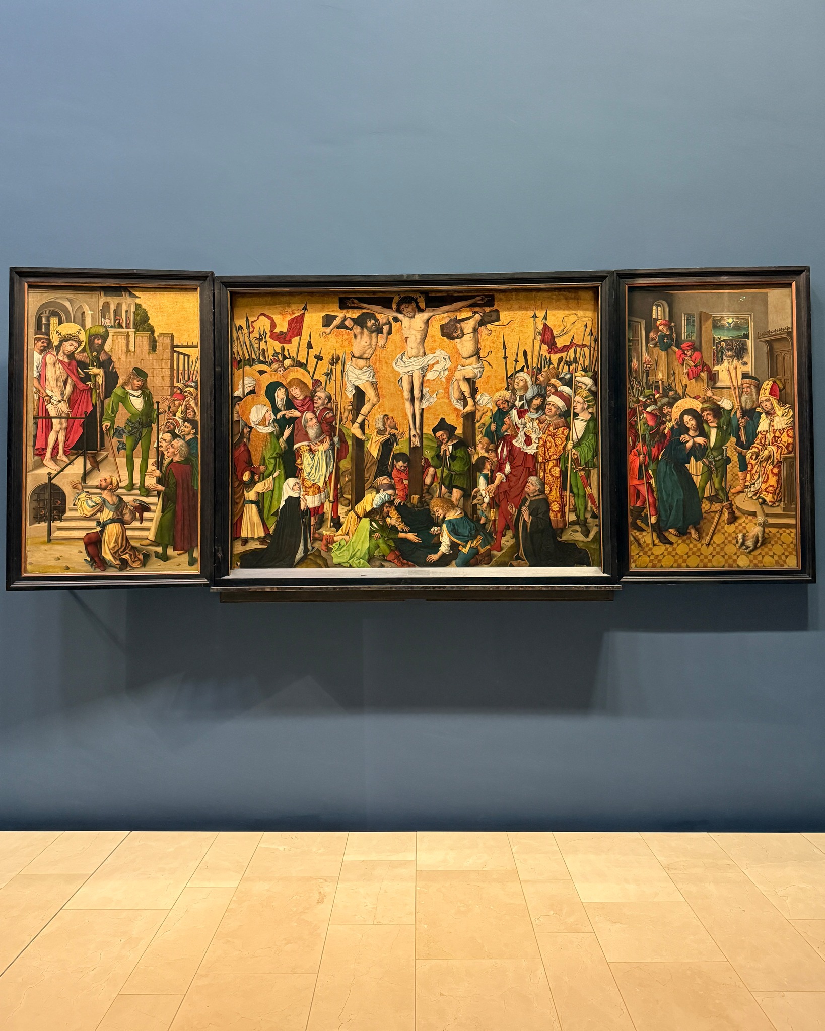 Ein dreiteiliges religiöses Gemälde hängt an einer blauen Wand. Links ist die Geißelung einer Figur zu sehen, in der Mitte die Kreuzigung mit drei Gekreuzigten und einer großen Menschenmenge, und rechts eine Trauergruppe um einen toten Körper. (KI-Alternativtext)