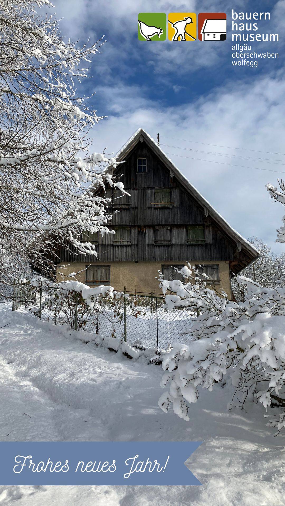 Winterliche Ansicht des Hauses Häusing. Verschneite Äste rahmen das dunkle Holzhaus mit steilem Dach. Ein Zaun und ein schneebedeckter Weg im Vordergrund. Blauer Himmel mit Wolken. Oben rechts das Museumslogo, unten der Schriftzug „Frohes neues Jahr!“.