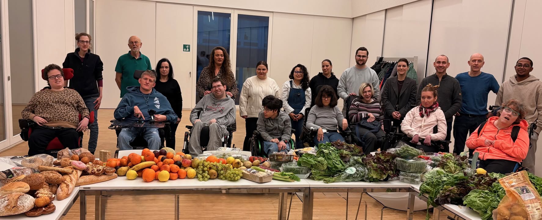 Dieser Alternativtext wurde KI-generiert: Eine Gruppe von Menschen steht für ein gemeinsames Foto in einem hellen Raum. Vor ihnen steht ein Tisch mit Brot, Obst und Gemüse.