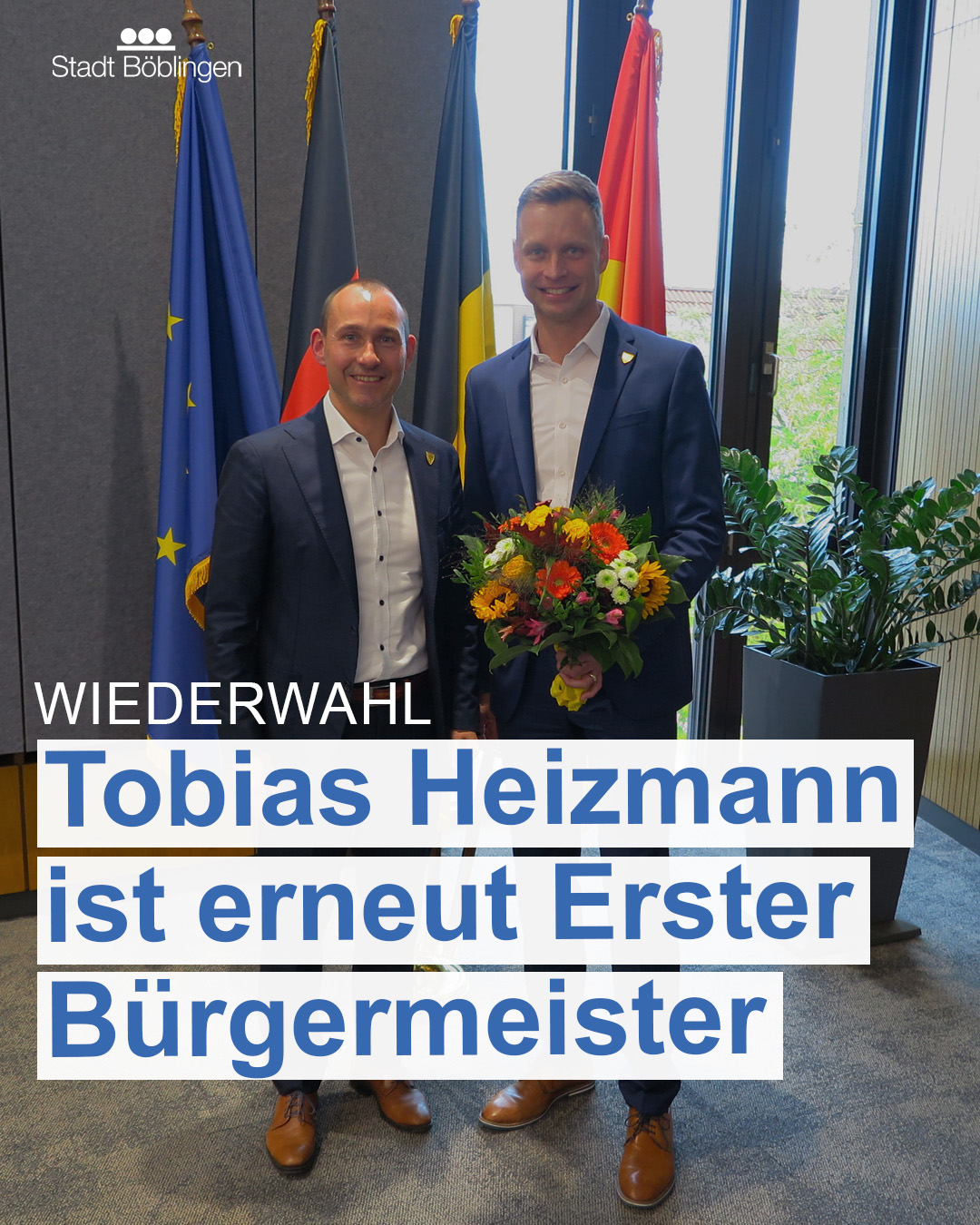 Dieser Alternativtext wurde KI-generiert: Zwei Personen stehen vor einer Flaggenwand und einer Pflanze. Im Vordergrund befindet sich ein Banner mit Aufschrift.