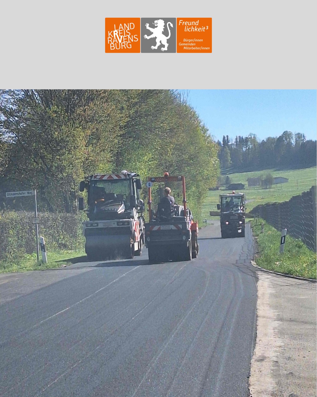 Dieser Alternativtext wurde KI-generiert: Eine asphaltierte Straße führt durch eine ländliche Landschaft. Mehrere landwirtschaftliche Fahrzeuge sind auf der Straße unterwegs.