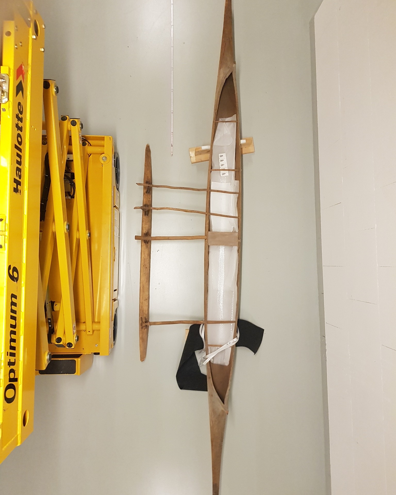 Dieser Alternativtext wurde KI-generiert: Ein schmales Boot, vermutlich ein Kajak, hängt an einer weißen Wand. Im Hintergrund ist ein Teil einer Hebebühne zu sehen.