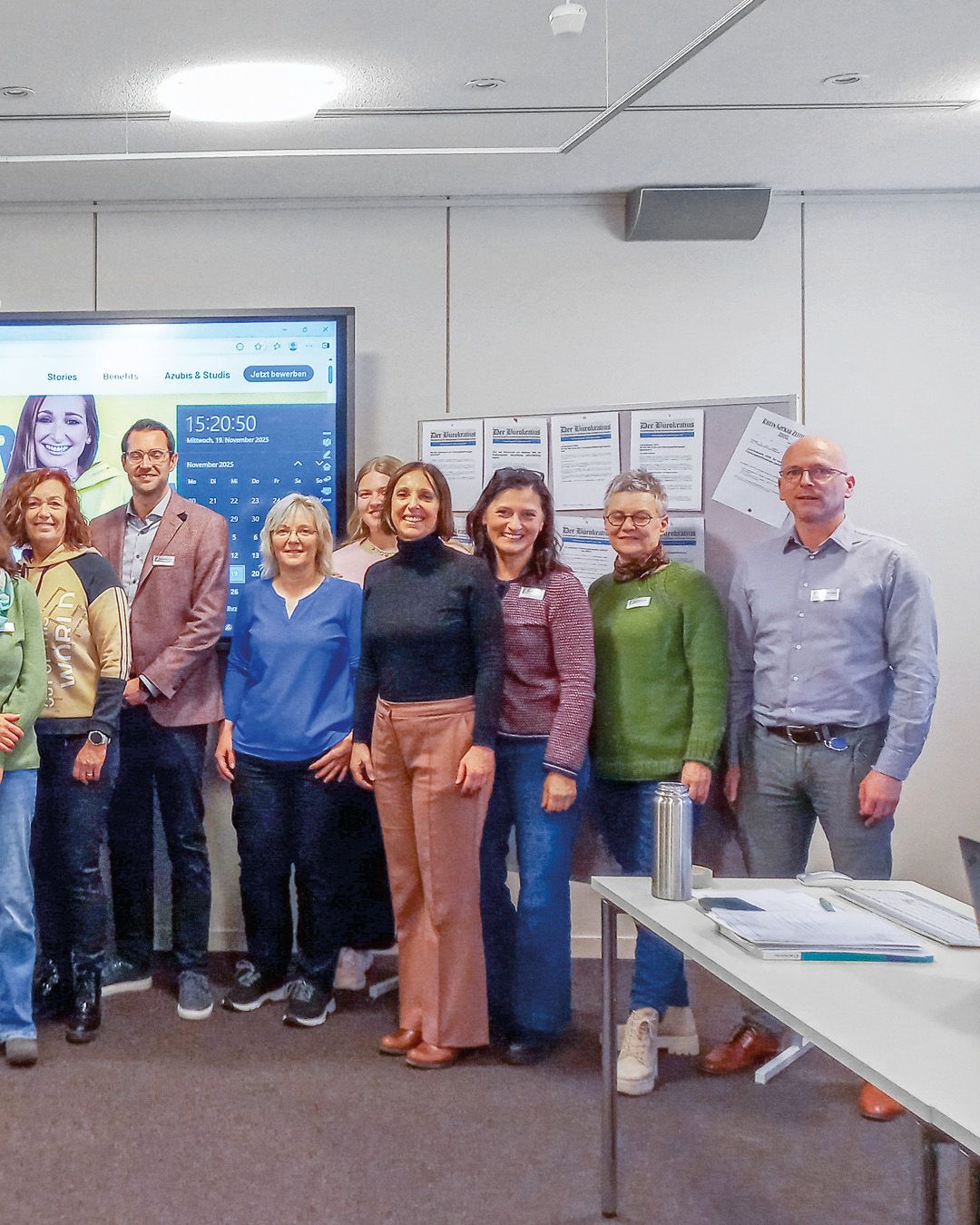 Dieser Alternativtext wurde KI-generiert: Eine Gruppe von Personen steht in einem Innenraum vor einer Wand mit Plakaten und einem Bildschirm. Ein Kalender ist im Hintergrund sichtbar.