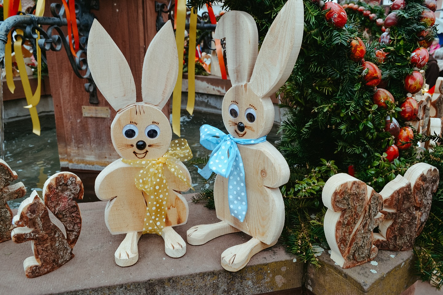 Zwei hölzerne Osterhasenfiguren mit Schleifen stehen neben einem Brunnen. Weitere hölzerne Figuren sind ebenfalls zu sehen.