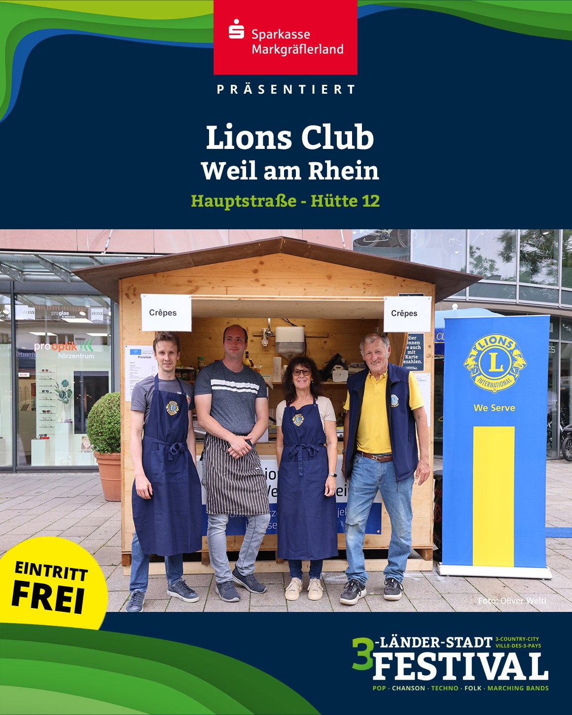 Eine farbige Aufnahme zeigt Personen vor einem Crêpes-Stand. Banner und Logos von Organisationen sind im Bild zu sehen.