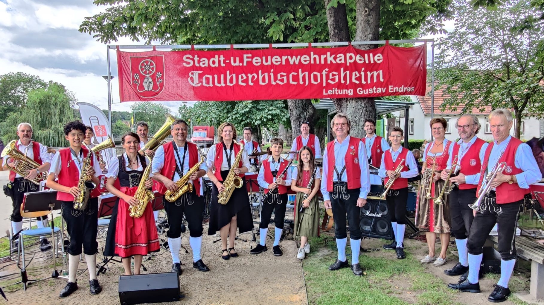 Eine Gruppe von Musikern in traditioneller Tracht mit roten Westen steht im Freien vor einem roten Banner, das "Stadt-u. Feuerwehrkapelle Tauberbischofsheim" und "Leitung Gustav Endres" beschriftet. Die Personen halten verschiedene Blasinstrumente wie Saxophone, Trompeten und ein Euphonium. (KI-Alternativtext)
