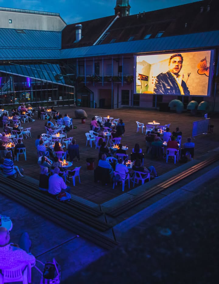 Open-Air-Kino in einem Innenhof bei Dämmerung. Personen sitzen an Tischen und auf Stufen und schauen auf eine große Leinwand an der Gebäudefassade, auf der eine Filmszene mit einer Person in einer Jacke zu sehen ist. Die Umgebung ist blau beleuchtet. (KI-Alternativtext)