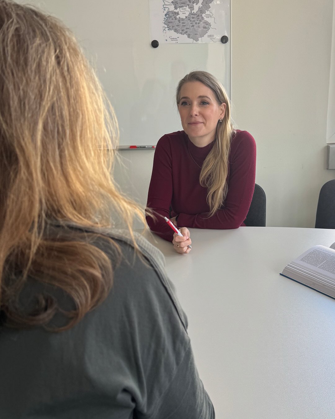 Dieser Alternativtext wurde KI-generiert: Zwei Personen sitzen sich an einem Tisch gegenüber. Im Hintergrund ist eine Weltkarte an einer weißen Wand zu sehen.