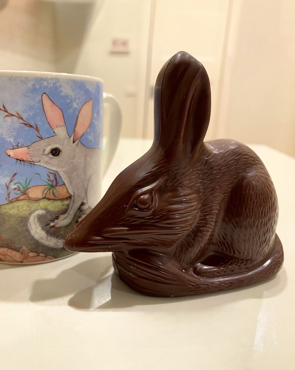 Dieser Alternativtext wurde KI-generiert: Eine Tasse mit einem Hasenmotiv steht neben einer Schokoladenhasenfigur. Das Bild ist farbig und zeigt die Objekte auf einer hellen Oberfläche.