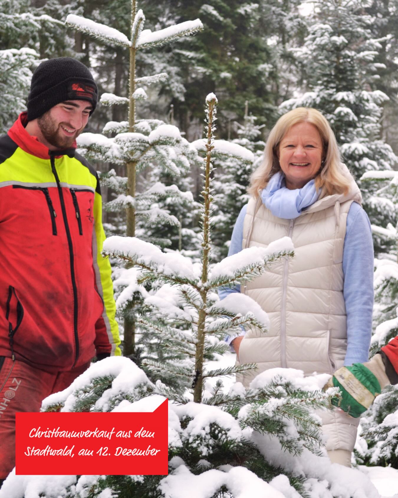 Dieser Alternativtext wurde KI-generiert: Zwei Personen stehen vor verschneiten Tannenbäumen. Im Vordergrund ist ein Schild mit einem Text zu sehen.
