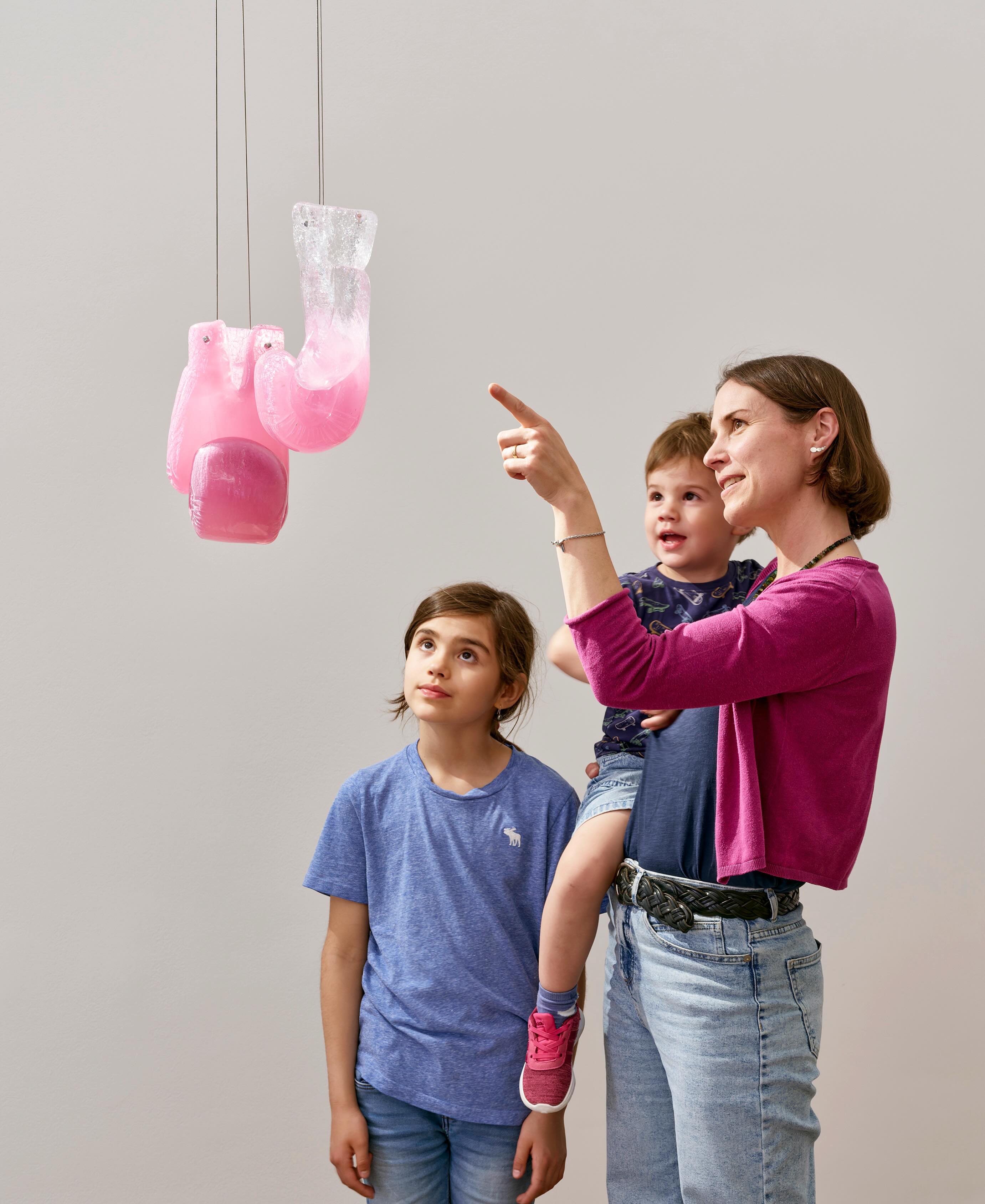 Dieser Alternativtext wurde KI-generiert: Eine Person zeigt auf hängende, rosa Skulpturen, während zwei weitere Personen aufmerksam beobachten. Das Bild ist farbig und zeigt eine Innenraumgestaltung.