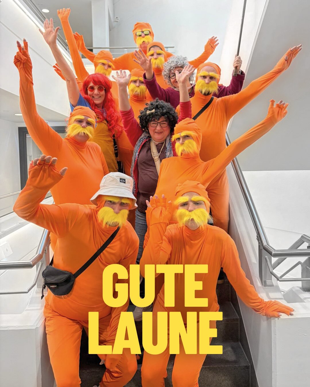 Dieser Alternativtext wurde KI-generiert: Eine Gruppe von Personen steht auf einer Treppe und trägt orangefarbene Kostüme mit Bart. Im Vordergrund ist ein Schild mit der Aufschrift "GUTE LAUNE" zu sehen.