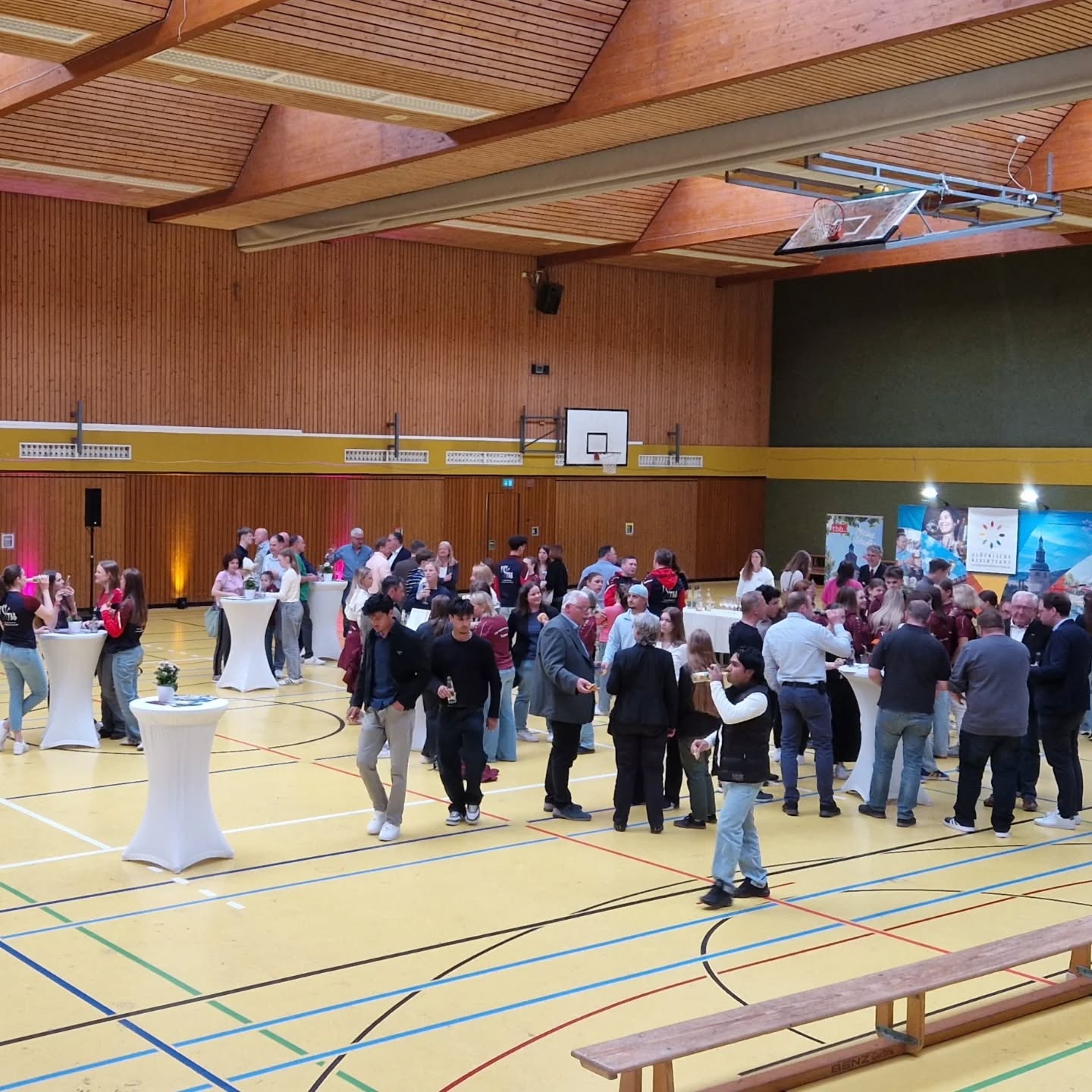 Dieser Alternativtext wurde KI-generiert: Eine Gruppe von Personen steht und unterhält sich in einer großen Sporthalle. Im Hintergrund sind Basketballkörbe und Fenster zu sehen.