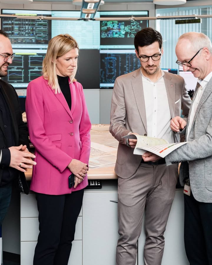 Dieser Alternativtext wurde KI-generiert: Mehrere Personen stehen in einem modernen Raum mit großen Bildschirmen und betrachten ein Dokument. Die Farbgebung ist überwiegend hell, mit einem auffälligen pinken Blazer.