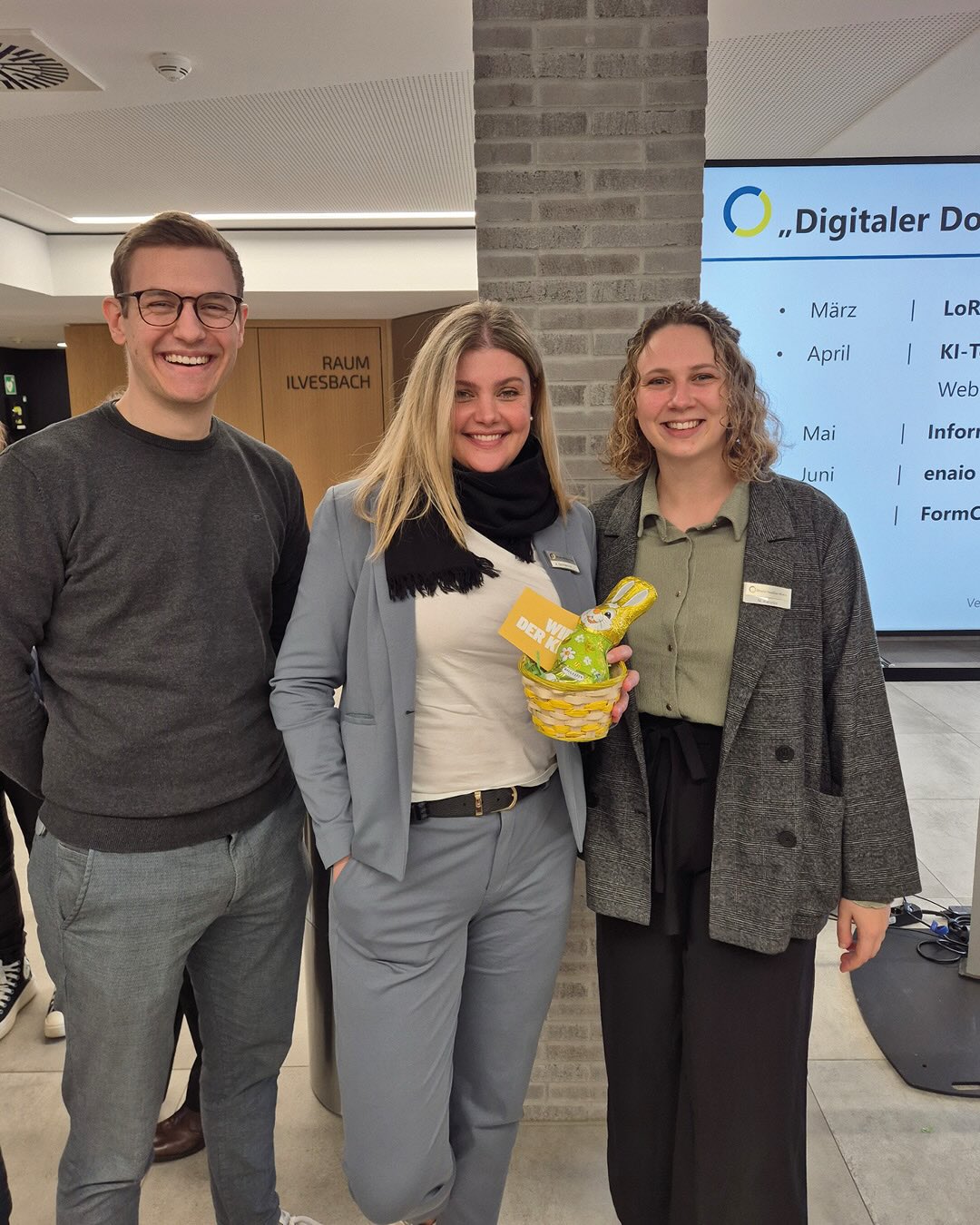 Dieser Alternativtext wurde KI-generiert: Drei lächelnde Personen stehen nebeneinander vor einer Backsteinwand und einem Whiteboard. Eine Person hält einen kleinen Korb.
