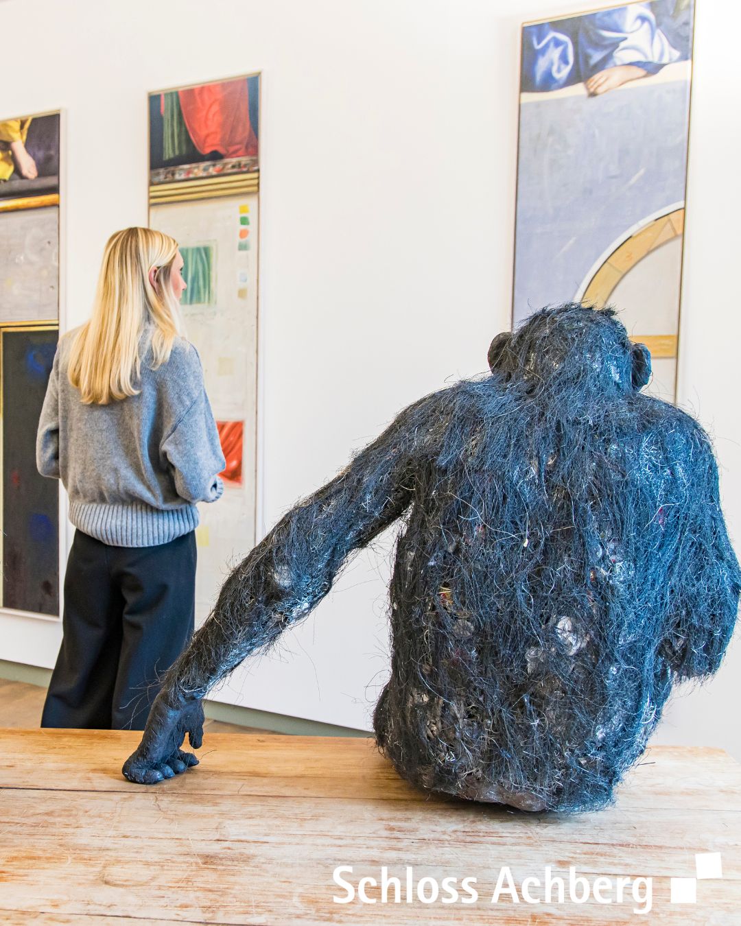 Eine Person betrachtet Kunstwerke in einem hellen Raum. Im Vordergrund steht eine blaue, behaarte Skulptur, die einem Affen ähnelt.