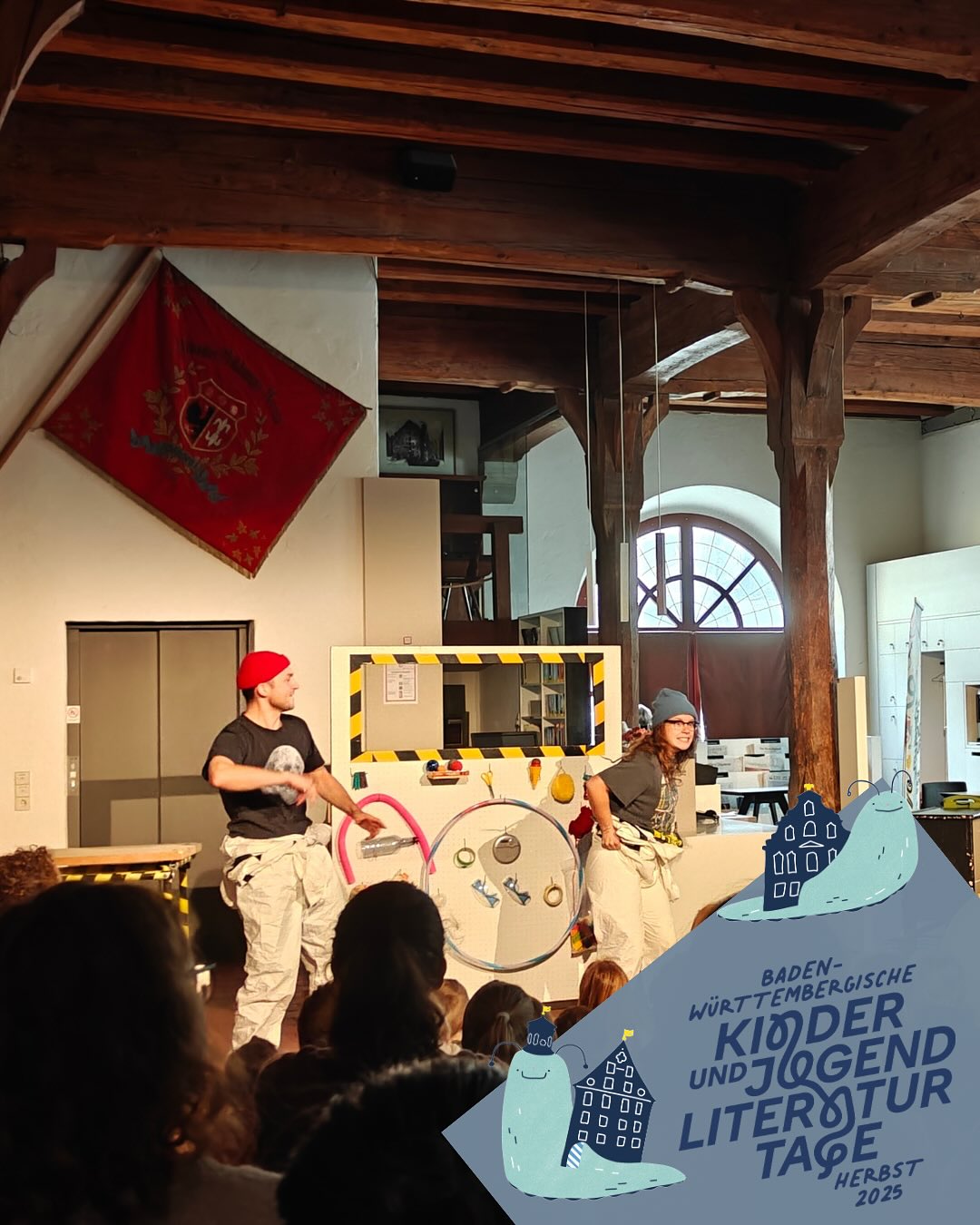 Dieser Alternativtext wurde KI-generiert: Ein Innenraum mit einer Bühne und Zuschauern. Auf der Bühne stehen Personen vor einem Kamin und einem Banner mit Text und Illustrationen.