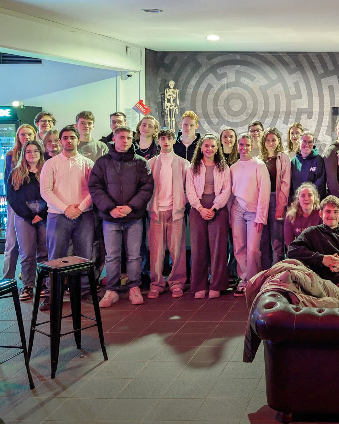 Dieser Alternativtext wurde KI-generiert: Eine Gruppe von Menschen steht in einem Raum mit einer kunstvollen Wanddekoration. Im Vordergrund befindet sich ein rotes Sofa und eine Person, die darauf sitzt.