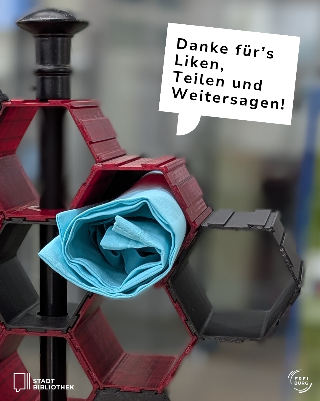 Dieser Alternativtext wurde KI-generiert: Ein farbiges Bild zeigt eine abstrakte Skulptur aus roten und schwarzen Bausteinen. Eine blaue Stoffblume ist darin integriert und ein Textfeld bedankt sich für Interaktionen.