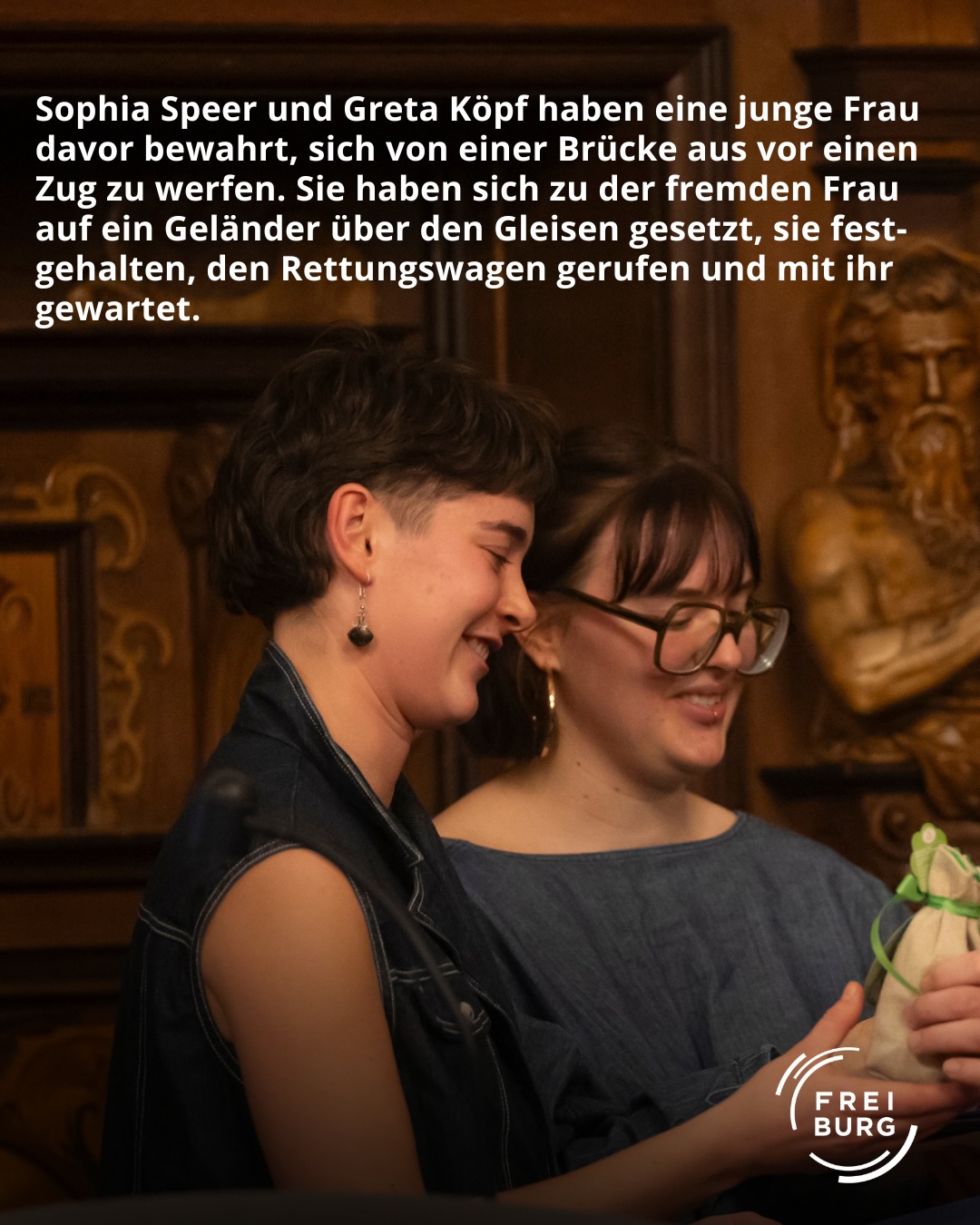 Dieser Alternativtext wurde KI-generiert: Schwarz-weißes Porträt von zwei Personen, die einander zugewandt sind. Im Hintergrund ist ein Bahnhofselement und eine Gleisanlage zu sehen.