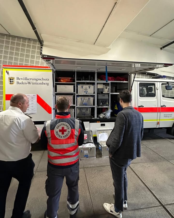 Dieser Alternativtext wurde KI-generiert: Ein weißer Rettungswagen mit roten Kreuzen ist in einem Innenraum abgebildet. Mehrere Personen stehen um das Fahrzeug und betrachten den Laderaum.