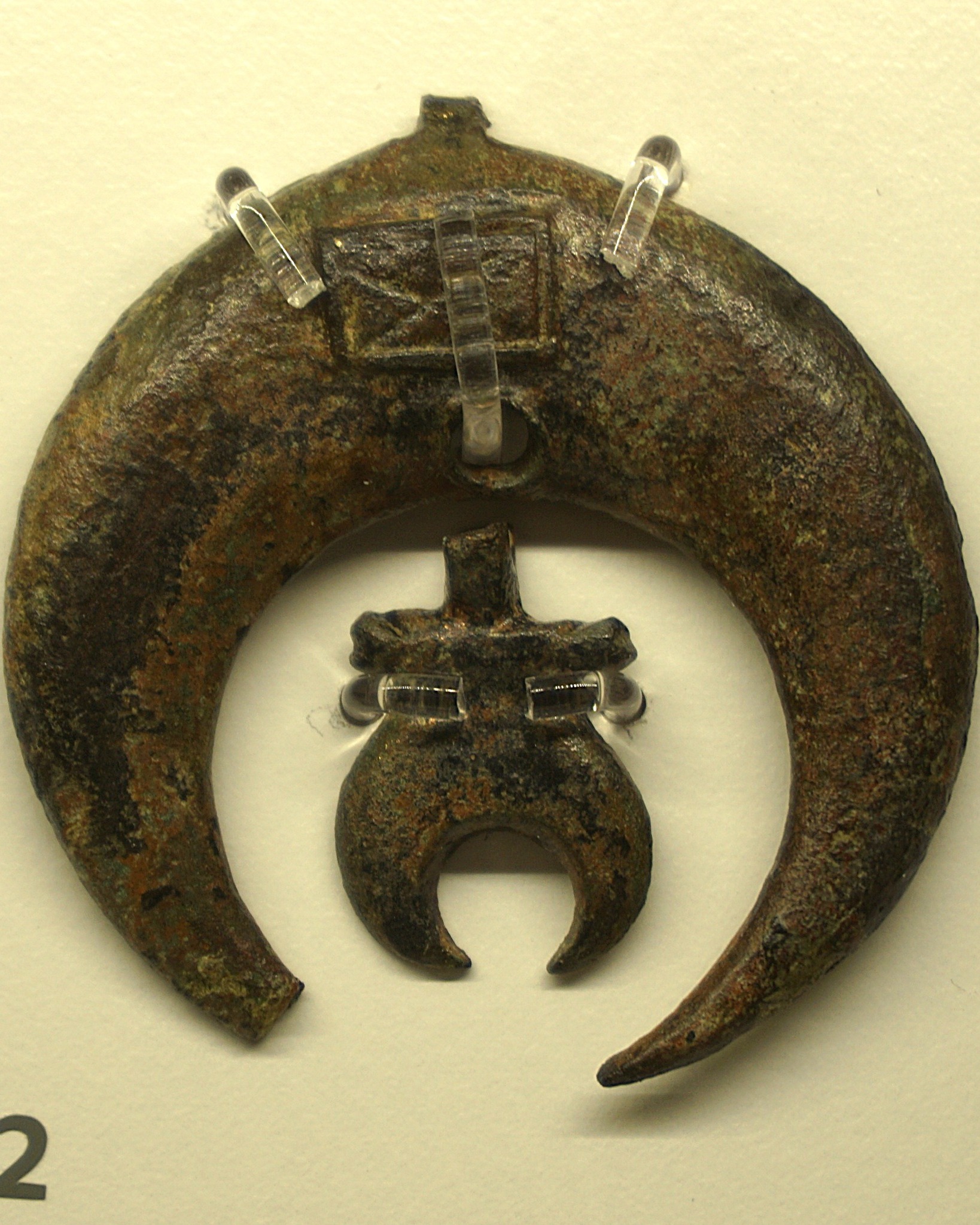 Dieser Alternativtext wurde KI-generiert: Ein dunkler, metallener Gegenstand in Form eines Halbmonds hängt an einer transparenten Aufhängung. Darunter befindet sich ein weiterer, tropfenförmiger metallener Anhänger.