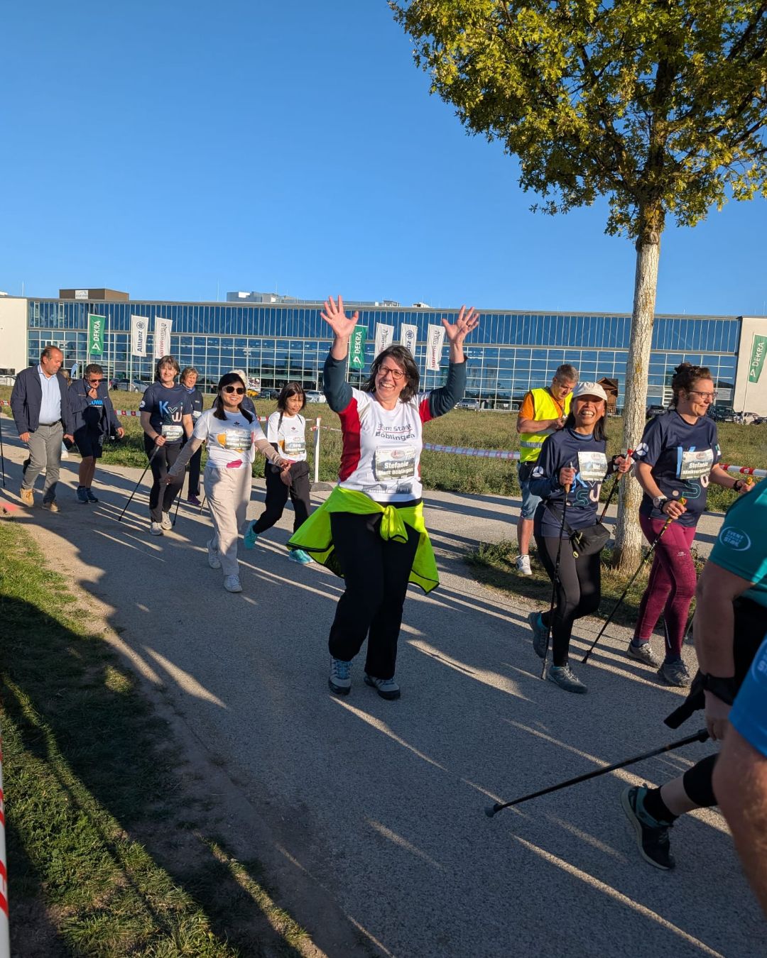 Eine Gruppe von Menschen läuft im Freien auf einem asphaltierten Weg. Die Person in der Mitte hebt beide Arme und lächelt in die Kamera. Sie trägt ein T-Shirt mit der Aufschrift „Team Stadt Boblingen“. Im Hintergrund steht ein großes Gebäude mit Werbeflaggen, darunter „DEKRA“ und „Allianz“. (KI-Alternativtext)