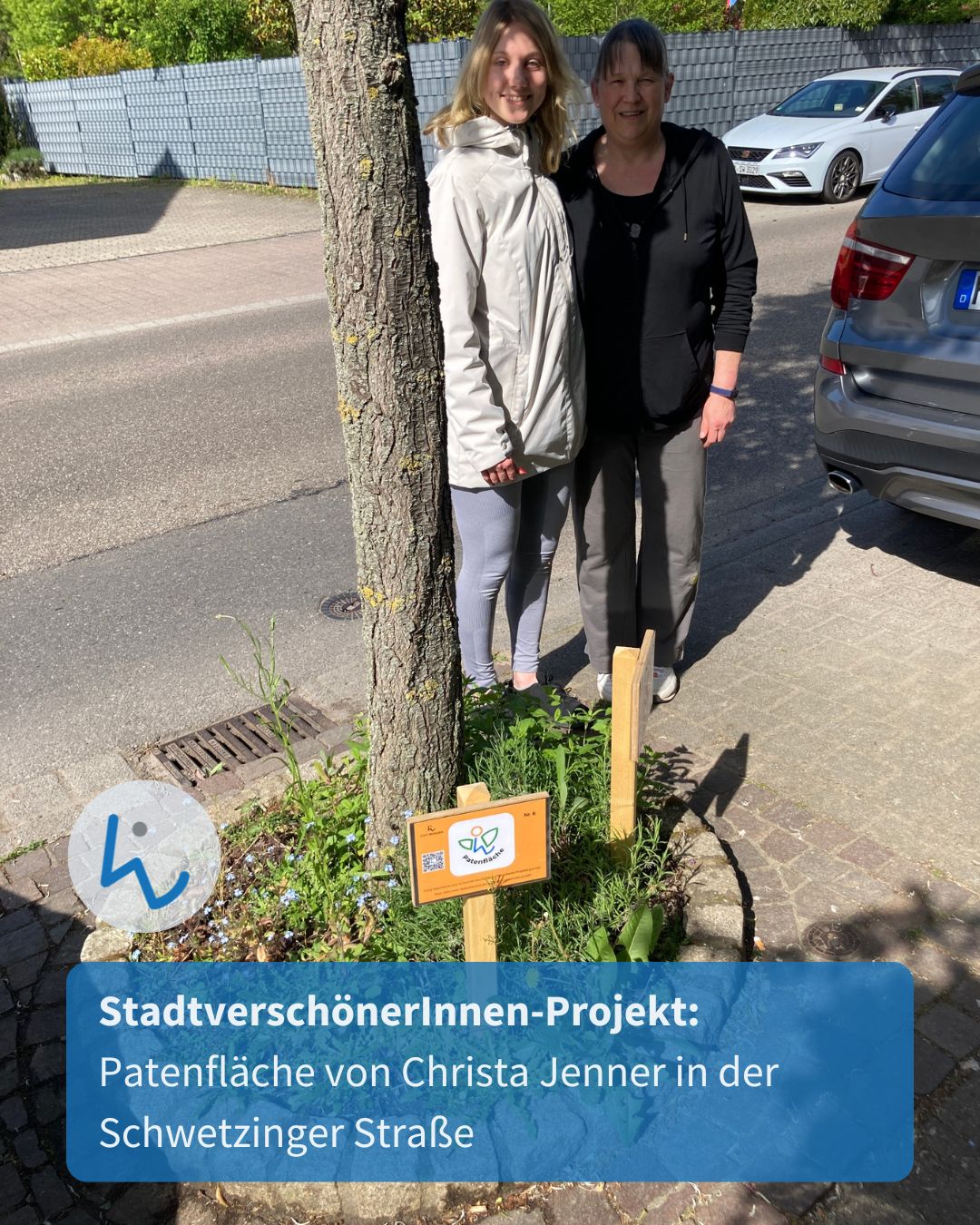 Dieser Alternativtext wurde KI-generiert: Zwei Personen stehen neben einem Baum und einem kleinen Beet. Im Hintergrund sind Autos und eine Mauer zu sehen.